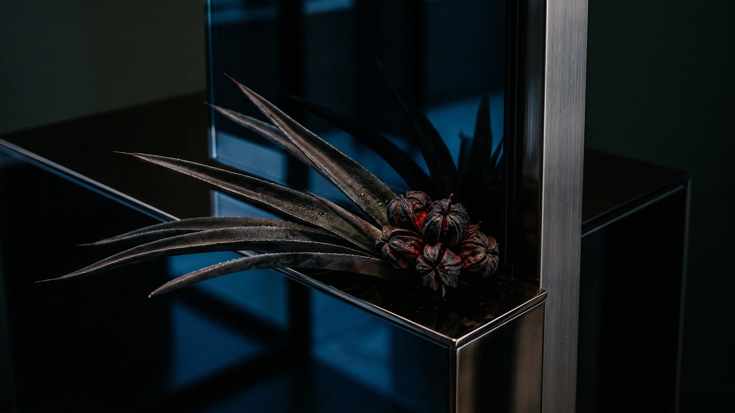 Long dark blades and a cluster of red and black seed pods at the foot of a steel post, with blue glass behind.